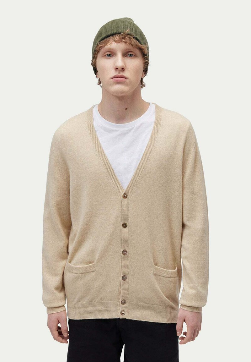 Beige kofta i mjukt material, med V-ringning, knapplist och två framfickor. Buren över en vit T-shirt.