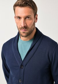 Námořnický modrý kardigán s šálovým límcem a třemi knoflíky, přes lehké tyrkysové tričko, s hladkou texturou a neformálním designem.