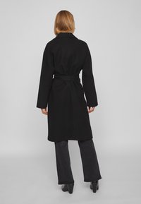 Γυναίκα με μαλλιά ξανθά μέχρι τους ώμους, φορώντας μαύρο παλτό με ζώνη, μαύρο παντελόνι και μαύρες μπότες με τακούνι, που στέκεται στραμμένη πλάτη σε απλό φόντο.