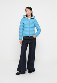 Lichtblauwe pufferjack met horizontale stiksels, een capuchon en ritssluiting. Gecombineerd met donkere wijde jeans en zwarte instapschoenen.