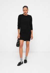 Vestido de suéter preto tricotado com mangas compridas e decote redondo, apresentando um detalhe de botão na lateral. Combinado com meias cinzas e sapatos pretos.