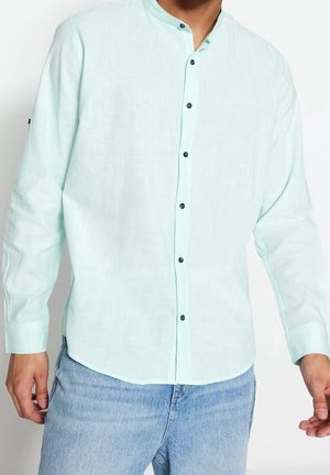 Man wearing light green mandarin collar long-sleeve shirt with black buttons and light blue jeans, standing against a white background.