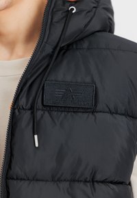 Melns puffa veste ar teksturētu virsmu, ar priekšējo rāvējslēdzēju un uzlīmes etiketē ar iebultu logo. Iekļauta aukla pielāgošanai.