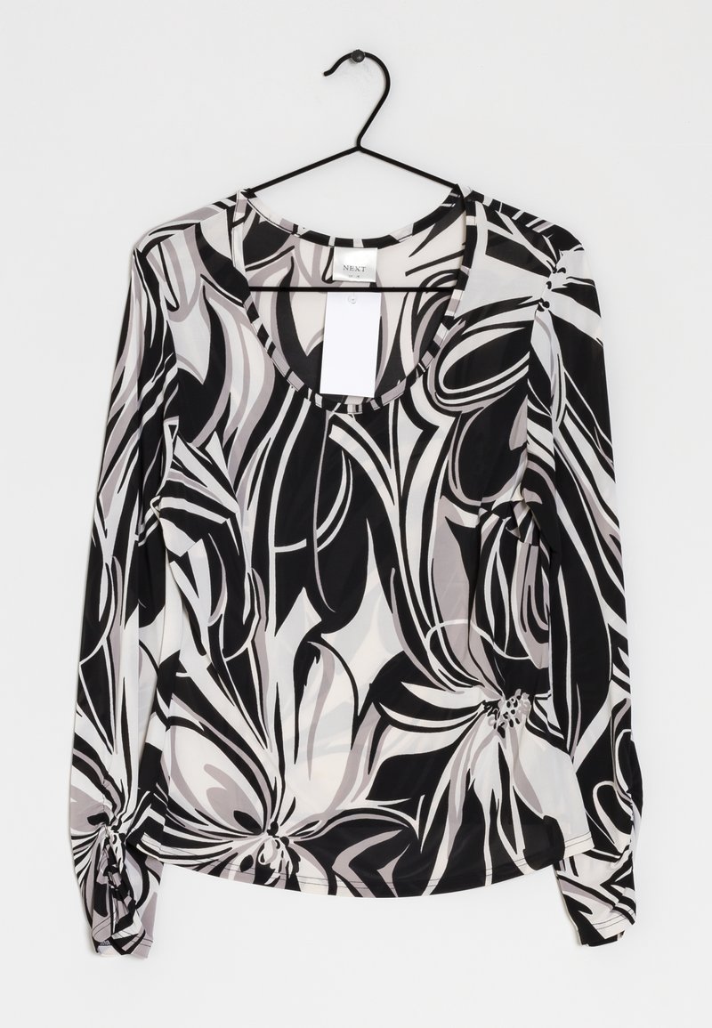 Blouse à manches longues avec motif floral noir, blanc et gris, col rond, suspendue sur un cintre noir contre un fond blanc.