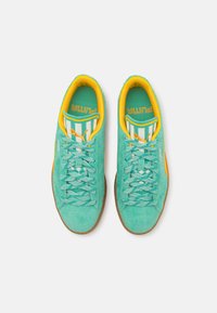 Tenisi din suede de un teal deschis, cu accente galbene, șireturi verzi cu model și o talpă din cauciuc texturată de culoare bej. Prezintă un design cu dungi pe limbă.