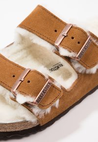 Brun mocka sandal med två justerbara remmar, prydd med kopparfjädrar och mjuk vit pälsfoder. Har en texturerad brun sula.