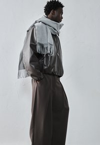 Moški v velikih svetlo rjavih hlačah, temno rjavem usnjenem jopiču in velikem svetlo sivošivem šalu stoji bočno ob strukturirani beli steni.