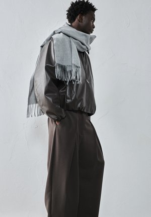 Muž v prehnane veľkých hnedých nohaviciach, tmavohnedej koženej bunde a veľkom svetlosivom šále, stojí bokom proti textúrovanej bielej stene.