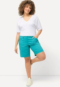 Camiseta blanca de cuello en V y pantalones cortos color teal con bolsillos, con una textura suave. El modelo está de pie de manera casual con una pierna doblada, llevando zapatillas blancas.