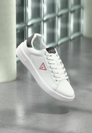 Białe niskie trampki Guess z czarnymi akcentami, sznurowadłami i czerwonym logo z boku, unoszące się nad polerowaną betonową podłogą w pobliżu ściany z bloków szklanych.