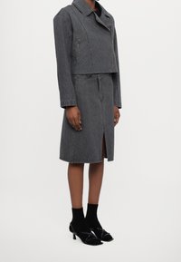 Giacca di jeans grigia dal design corto con cerniera, abbinata a una gonna di jeans grigia coordinata con spacco frontale. Indossati sandali neri con tacco.