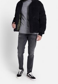 Schwarze Kord-Pufferjacke mit olivgrünem Innenfutter, grauem Strickpullover, grauen Jeans und schwarz-weißen Sneakers.