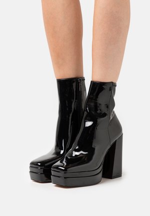 Bottes noires vernies à plateforme et talons carrés épais portées sur des jambes nues sur fond blanc uni.