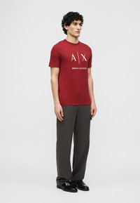 Άνδρας που στέκεται φορώντας κόκκινο T-shirt Armani Exchange, σκούρο γκρι φαρδιά παντελόνια και μαύρα επίσημα παπούτσια, μπροστά σε απλό ανοιχτό φόντο.