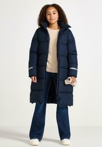 Cappotto puffer blu scuro con design lungo, caratterizzato da un colletto alto, cuciture orizzontali e dettagli riflettenti sulle maniche. Indossato su un maglione beige.