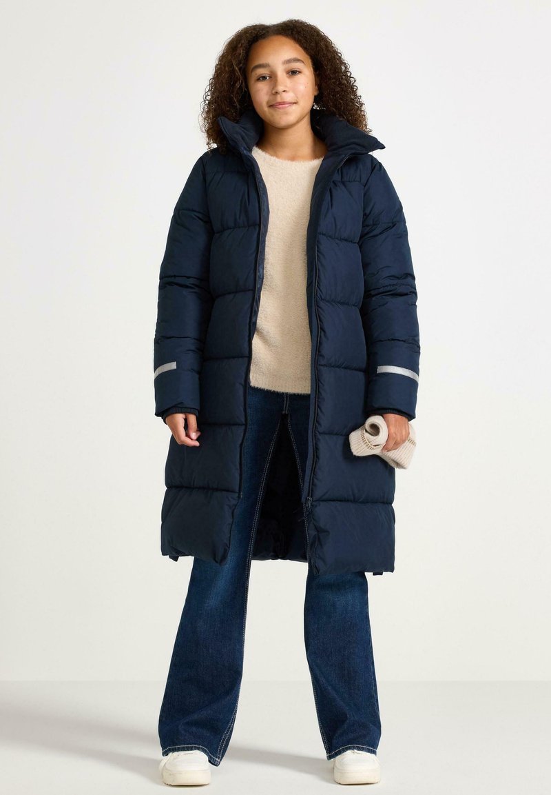 Cappotto puffer blu scuro con design lungo, caratterizzato da un colletto alto, cuciture orizzontali e dettagli riflettenti sulle maniche. Indossato su un maglione beige.