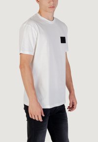 Camiseta blanca de manga corta de algodón, con un bolsillo rectangular negro en el lado izquierdo del pecho, combinada con jeans oscuros.