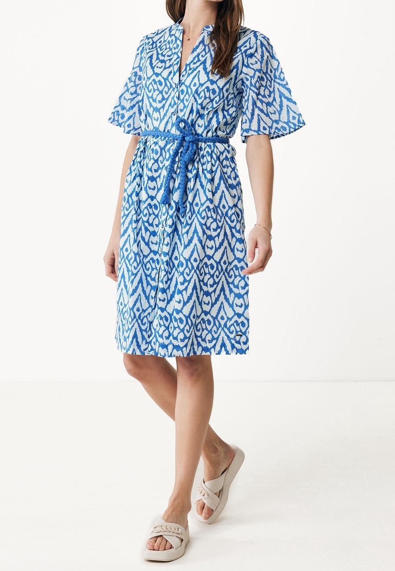 Femme portant une robe bleu et blanc à motifs longueur genoux, manches courtes, ceinture corde nouée, associée à des sandales slides beiges.