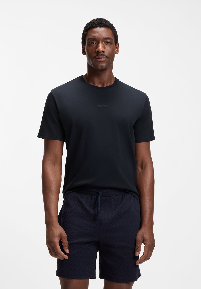 T-shirt noir à manches courtes et short sombre à motifs portés par un homme debout, les mains détendues le long du corps, sur fond blanc.