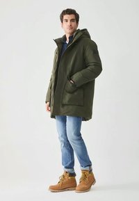 Manteau matelassé vert olive avec capuche, poches avant et détails noirs. Associé à un jean bleu clair et des bottes à lacets couleur fauve.