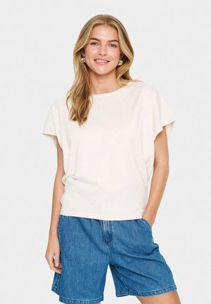 Top corto de manga corta color beige claro con un ajuste holgado y escote redondo, combinado con pantalones cortos de mezclilla azules. Tejido suave y texturizado. Diseño simple.