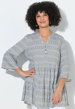 Vrouw met krullend haar die een grijze, patroonlange mouw tuniek draagt, met één hand op de heup, staand tegen een witte achtergrond.