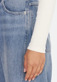 Jeans in denim azzurro chiaro con una tasca e un bottone argentato. Una camicia bianca a maniche lunghe è parzialmente visibile, con una texture a coste sulla manica.