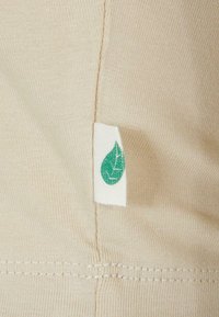 Beige fabric with a smooth texture, featuring a white tag with a green leaf design, stitched at the seam.