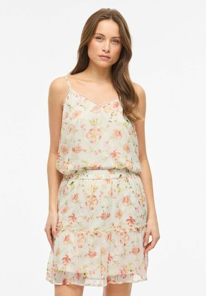 Frau trägt ein ärmelloses weißes Kleid mit rosa Blumenmuster, elastischer Taille und Rüschensaum, steht vor einem schlichten weißen Hintergrund.