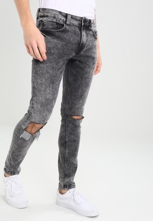 Mężczyzna w wyblakłych, czarnych, podartych obcisłych dżinsach, białych trampkach i białej koszuli na jednolitym tle.