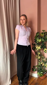 Νέα γυναίκα με ξανθά μαλλιά, που φοράει ανοιχτό μωβ καρό μπλούζα και μαύρο παντελόνι, στέκεται κοντά σε μια γλάστρα με φυτό μπροστά σε ένα ροζ τοίχο και μπεζ κουρτίνα.