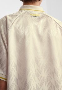 Ljust beige pikétröja med ett subtilt geometriskt mönster, korta ärmar och en kontrasterande gul krage. Märkeslogotyp syns på baksidan.