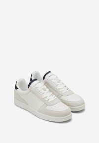 Paire de baskets basses blanches avec bout perforé, accents en daim, lacets et détail noir au talon et à la languette sur fond blanc.