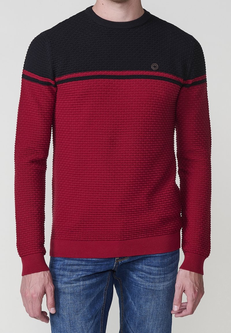 Homme portant un pull à manches longues texturé avec une partie supérieure noire et une partie inférieure rouge, associé à un jean bleu.