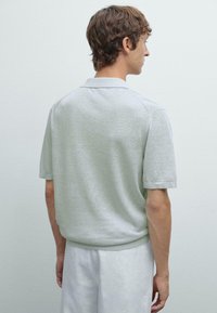 Světle šedé polo tričko s krátkým rukávem, klasickým límečkem, žebrovanými manžetami a hladkou strukturou. Zadní strana má minimalistické designové detaily.