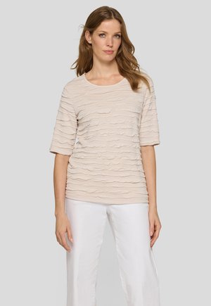Woman with light brown hair wearing a beige textured short-sleeve top and white pants standing against a plain light gray background.