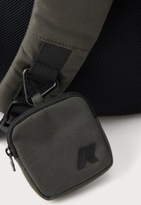 Tamnozelena platnena torba s naramenicom od mrežastog materijala, koja sadrži mali zippered toplić s mat crnim logotipom i teksturiranom površinom.
