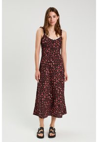 Robe noire à fleurs avec des fines bretelles, présentant un corsage ajusté et une jupe fluide, ornée de motifs de fleurs roses et rouges. Sandales noires.
