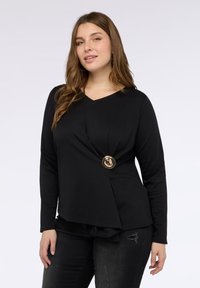 Maglietta nera a maniche lunghe con scollatura a V, caratterizzata da un design attorcigliato in vita e un bottone dorato sul lato sinistro. Tessuto morbido.