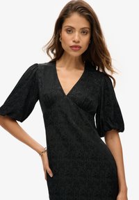 Vestido negro con escote en V y mangas abullonadas, que presenta un patrón texturizado. La modelo lleva pendientes dorados y muestra una pulsera en la muñeca derecha.