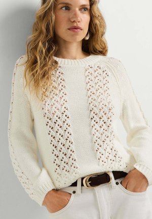 Mujer con cabello ondulado que lleva un suéter blanco de punto con patrones de encaje y pantalones de color claro, manos en los bolsillos, mirando ligeramente hacia un lado.