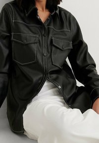 Svart läder skjortjacka med dubbla bröstfickor och kontrasterande vit sömnad. Designad med knäppning framtill och en avslappnad passform.