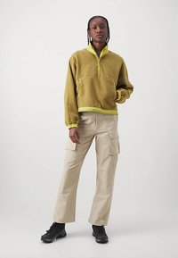 Πράσινη φλις μπλούζα ελιάς με μισό φερμουάρ, κίτρινες λεπτομέρειες και εμπρόσθια τσέπη· συνδυασμένη με μπεζ παντελόνι cargo και μαύρα αθλητικά παπούτσια.