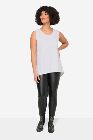 Donna con capelli ricci che indossa un top bianco senza maniche, leggings in pelle nera e stivaletti neri con borchie, in piedi con una mano sui fianchi.