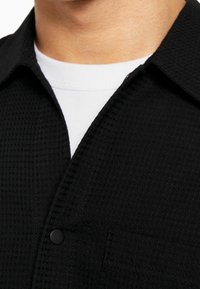 Svart texturerad skjorta med kragdesign, som har knappstängning och en ficka. Vit undertröja synlig under kragen.