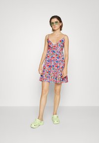 Vestido de verão com padrão floral e alças finas, apresentando tons de rosa, azul e verde. Combinado com tamancos verdes claros.