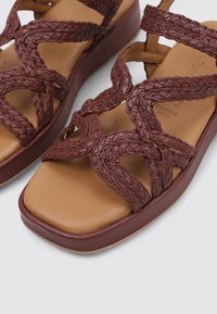 Sandale tissée marron avec des brides tressées, une semelle intérieure contour beige et une épaisse semelle bordeaux. Le design met l'accent sur le confort et la stabilité.