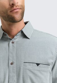 Primo piano della parte inferiore del viso di un uomo e di una camicia a disegni con colletto, bottoni e una tasca sul petto con dettagli scuri.