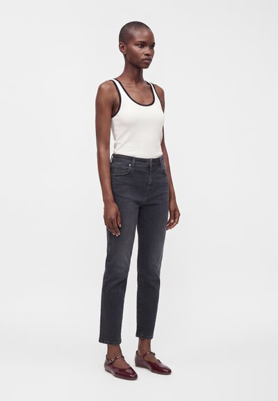 Femme debout portant un débardeur blanc avec une bordure noire, un jean gris foncé et des chaussures Mary Jane bordeaux sur un fond clair uni.