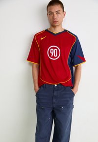 Jersey corto rojo y marino con ribete amarillo, que presenta un logotipo blanco circular "90". Emparejado con pantalones azules que tienen bolsillos y un corte relajado.
